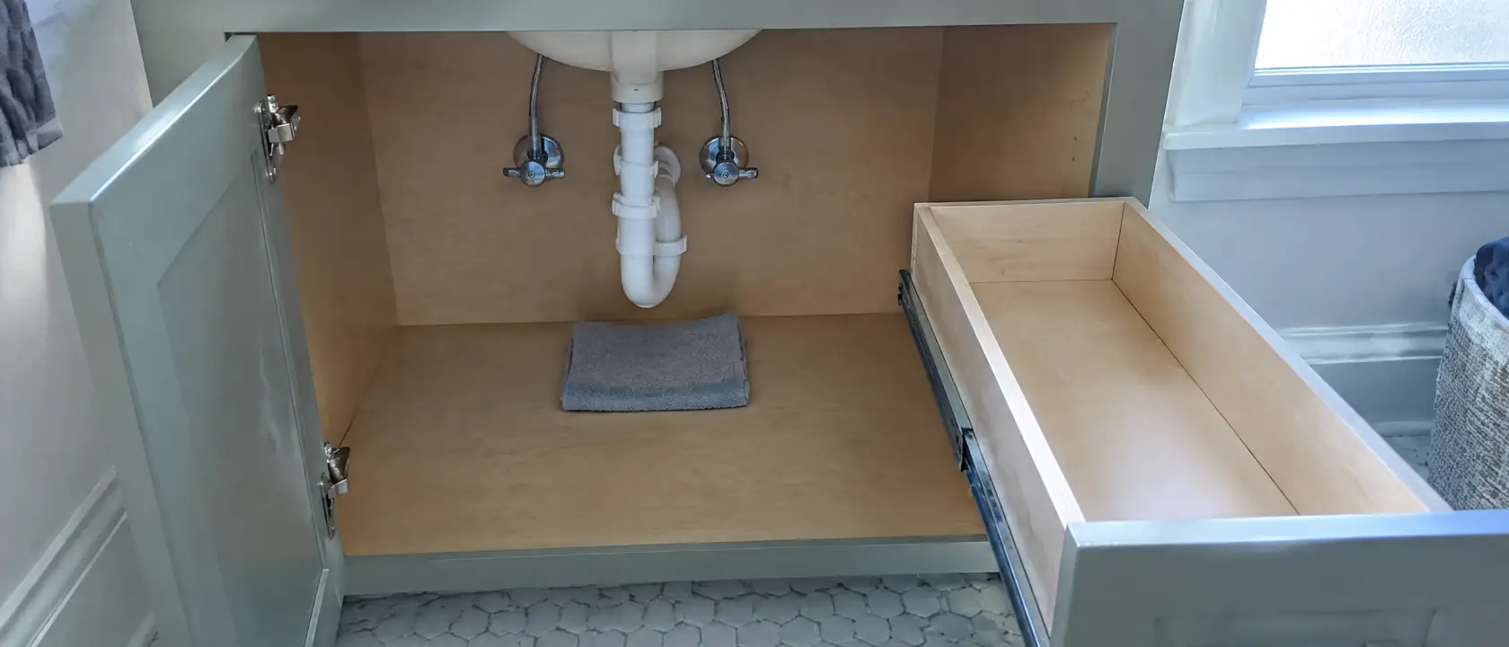Open vanity base showing the drain trap and shutoff valves over a dry plywood floor, the drawer pulled out on its runners