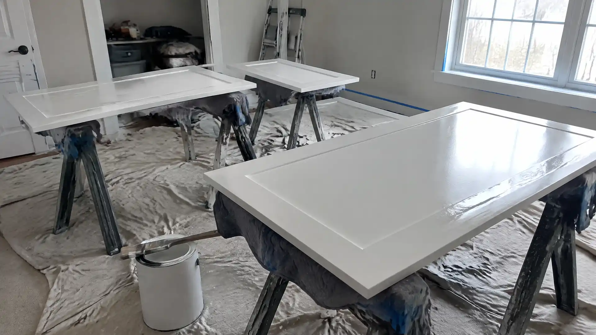 Freshly painted soft white cabinet doors drying on padded sawhorses over drop cloths