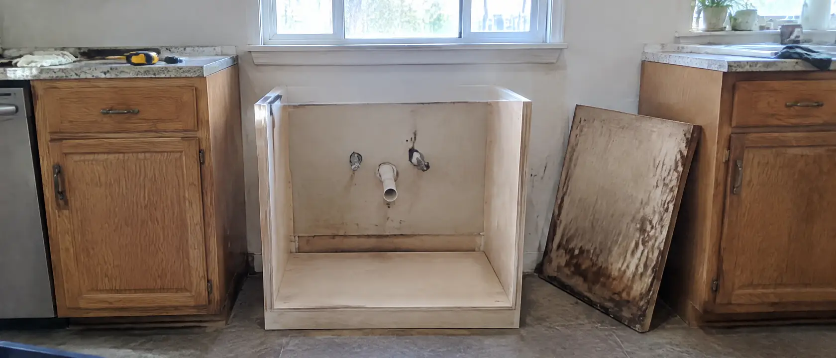 Kitchen run with the swollen old sink base removed and a new plywood box sliding in between two original cabinets