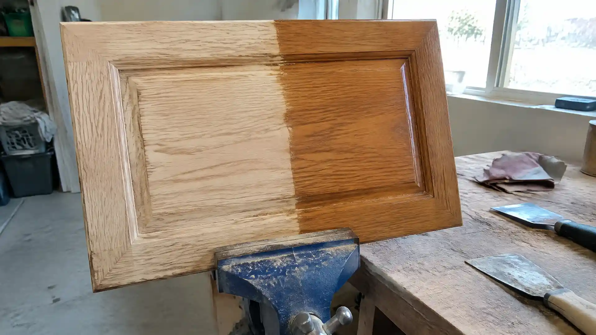 An oak cabinet door in a bench vise, half stripped to raw wood and half under old amber varnish
