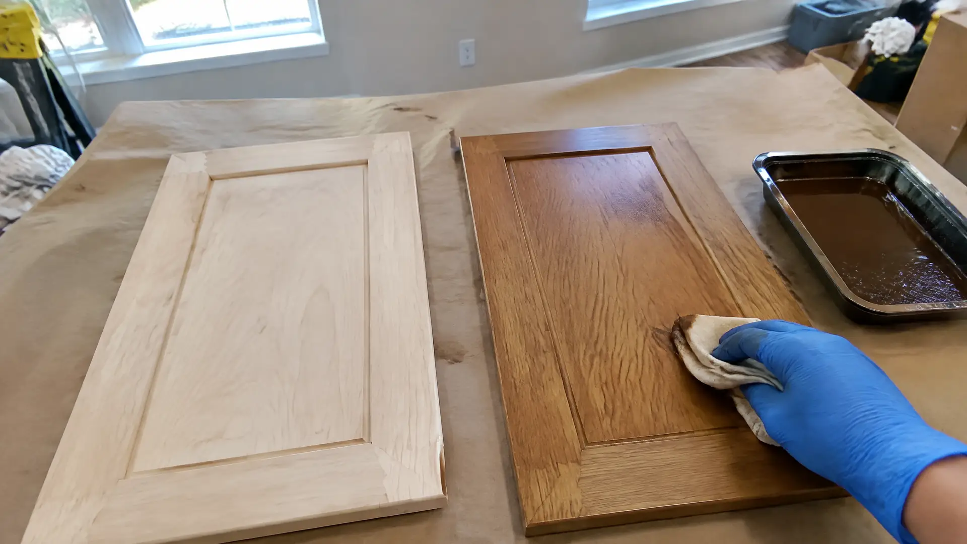 A gloved hand wiping warm brown stain along the grain of a maple cabinet door front
