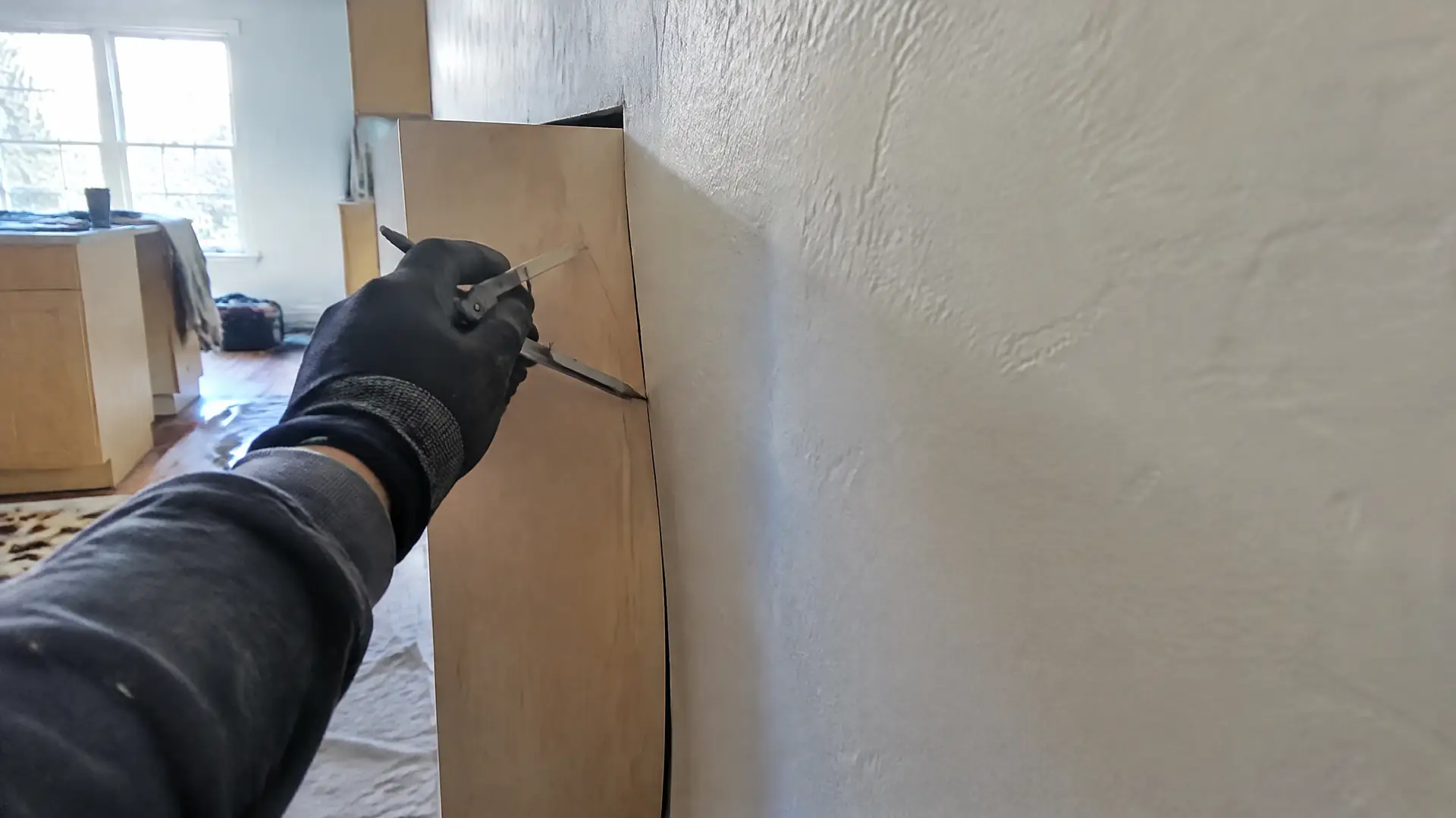A gloved hand running a compass down a cabinet end panel to scribe a bowed plaster wall