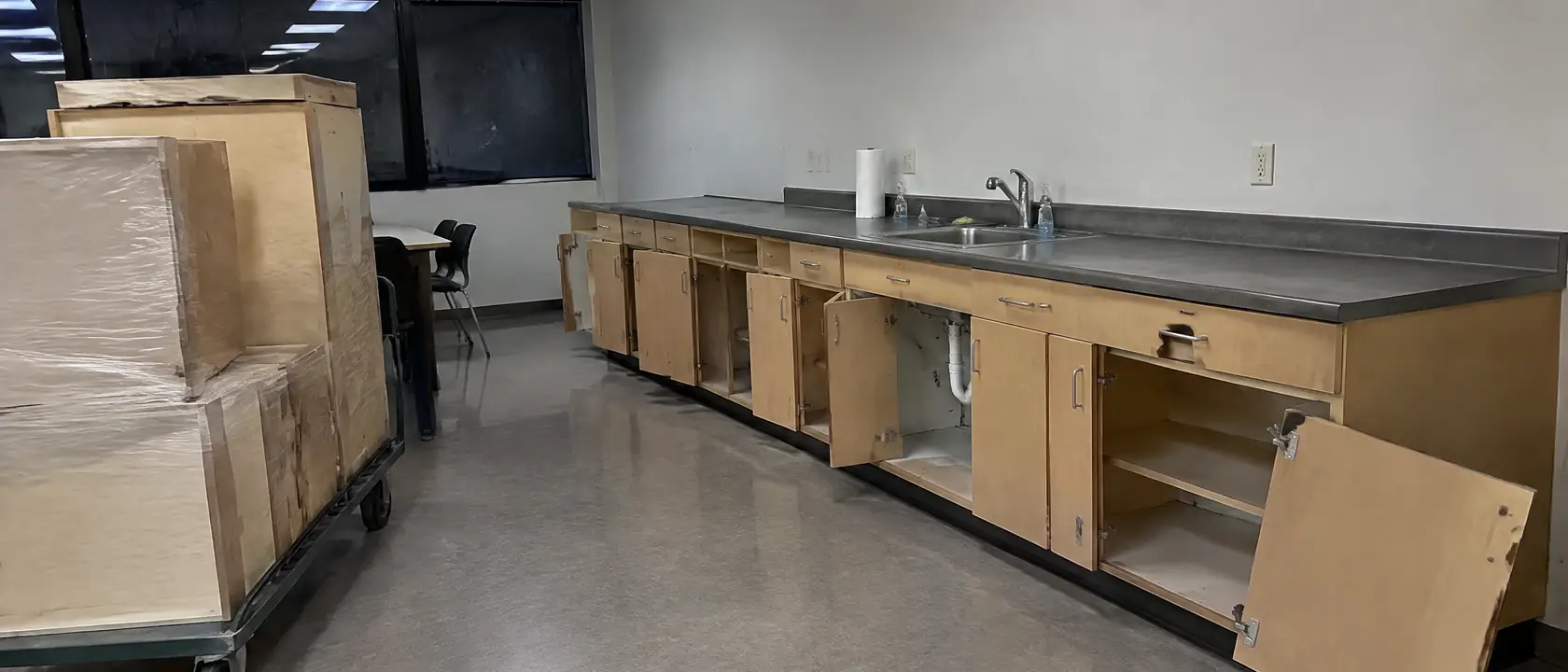 Office breakroom after hours, a worn laminate cabinet door sagging off its hinge, new wrapped cabinet boxes on a cart
