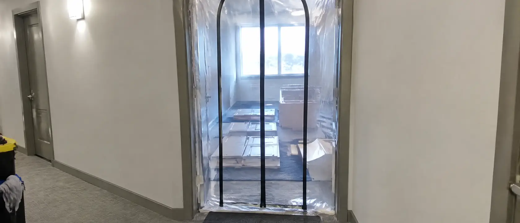 Condo doorway sealed with plastic sheeting and a zippered opening off a shared hallway, cabinet parts laid flat inside