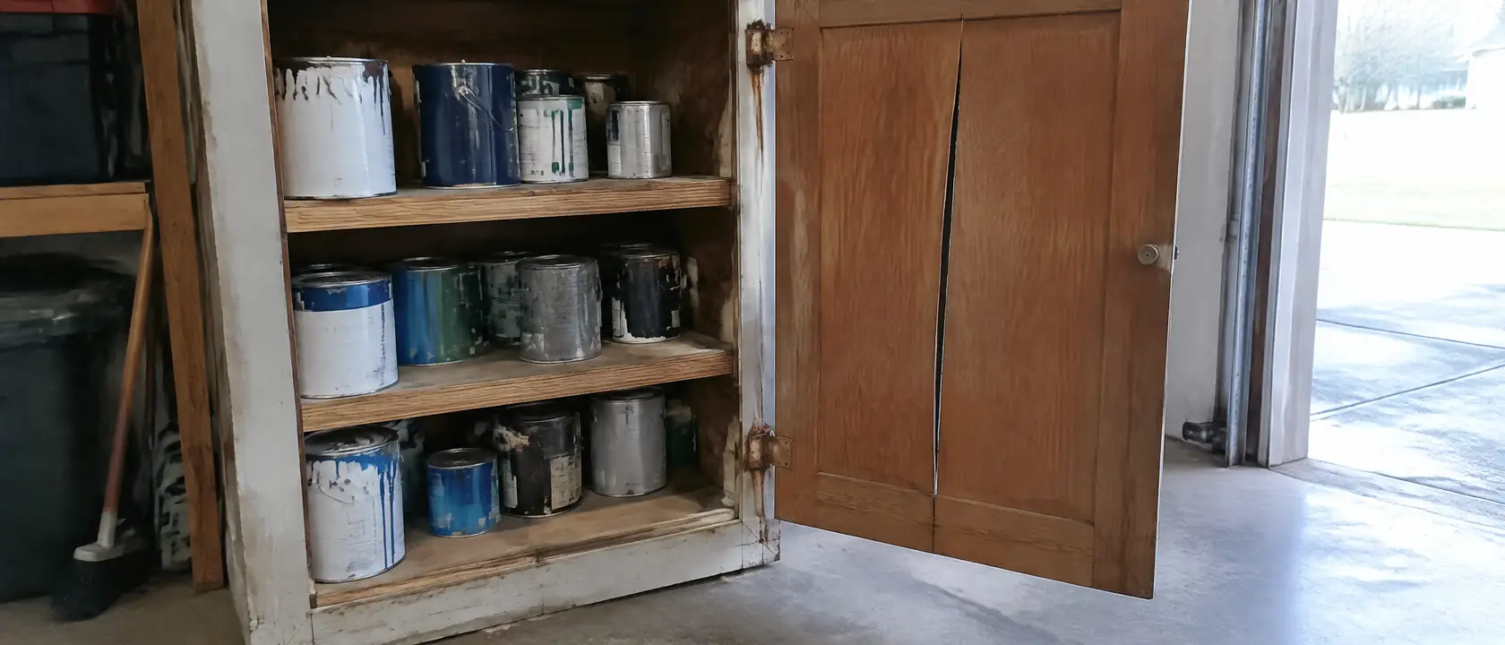 Garage cabinet door with its solid center panel split down the middle beside short, thick shelves of unlabeled paint cans
