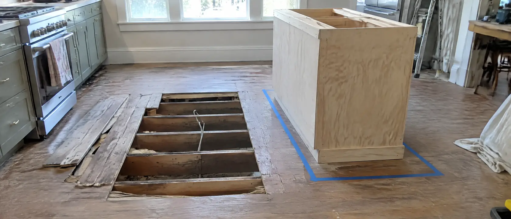 Kitchen floorboards lifted where an island will stand, a power cable coming up through the subfloor beside a plywood base