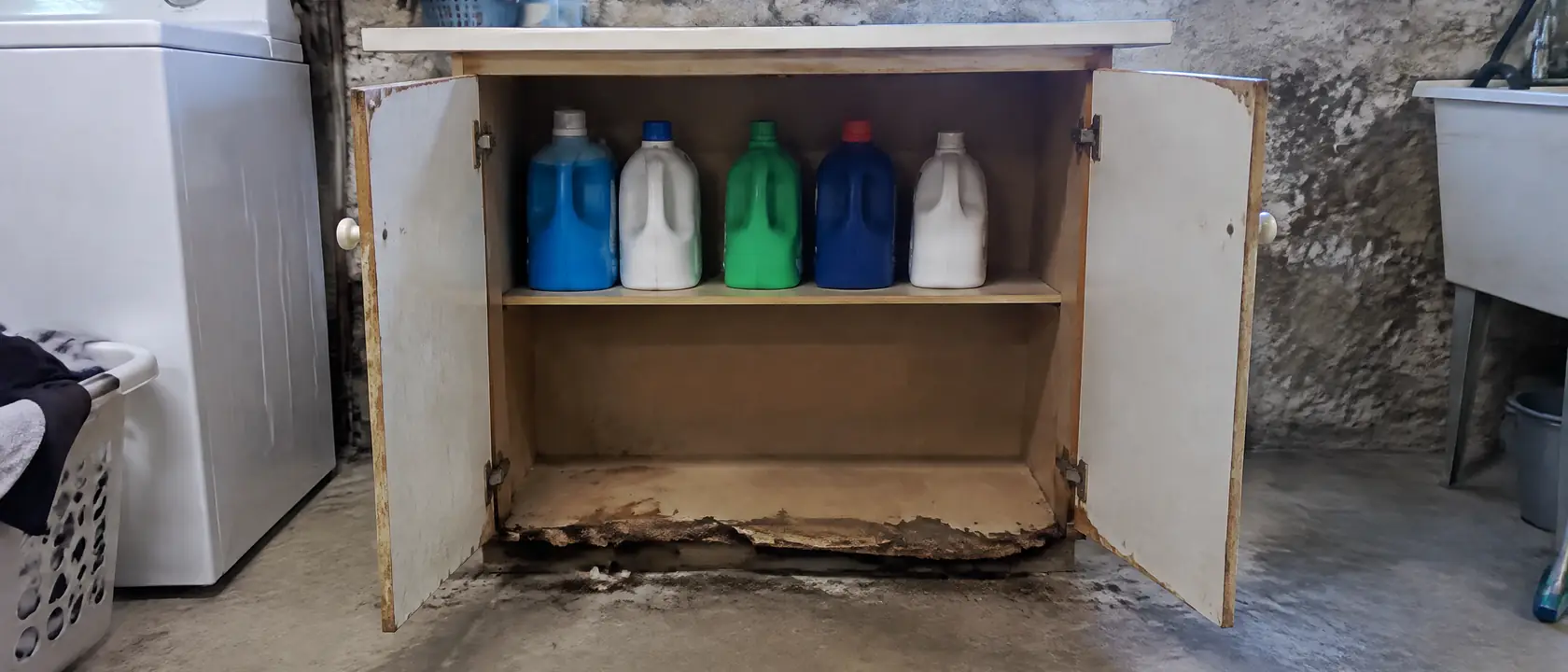 Old particleboard laundry cabinet swollen and crumbling at its bottom edge in a damp basement, detergent jugs on the shelf
