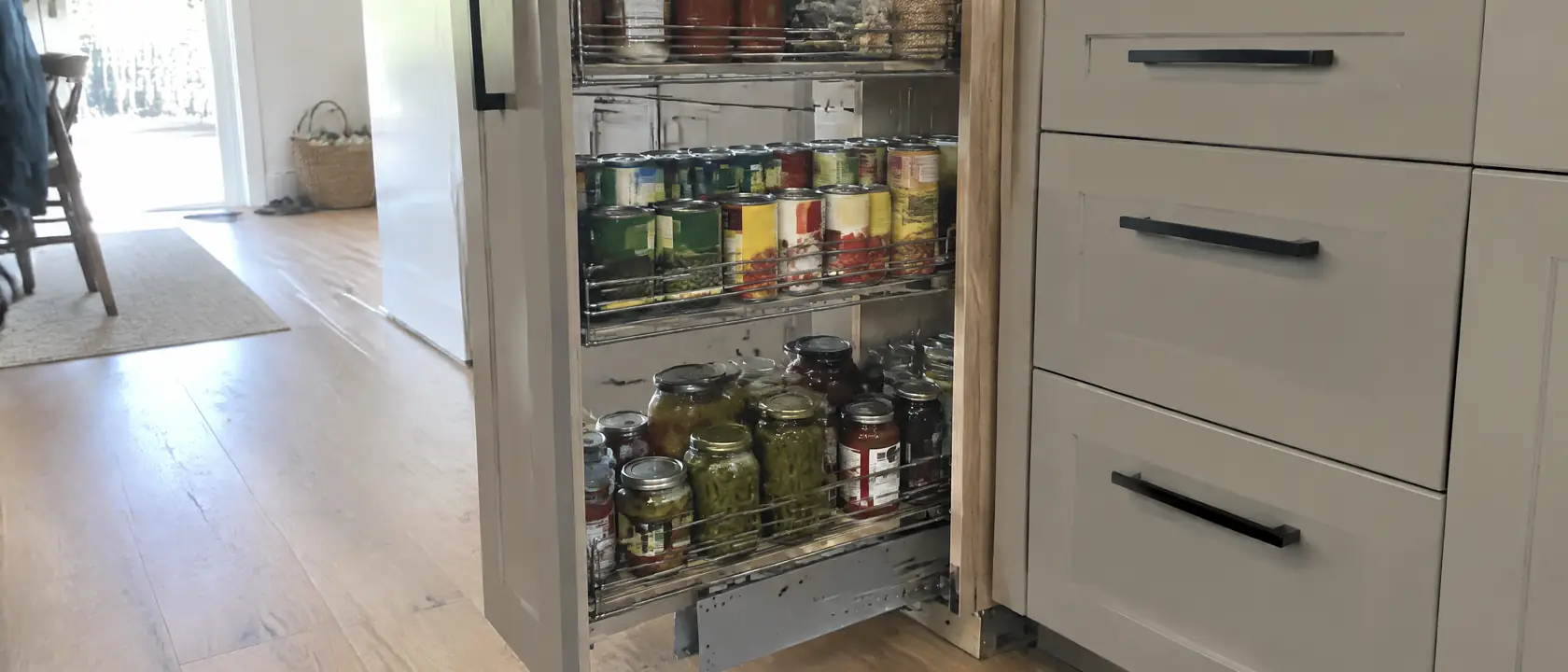 Tall pull-out pantry drawn open near the floor, its heavy steel runner carrying wire shelves of unlabeled cans and jars