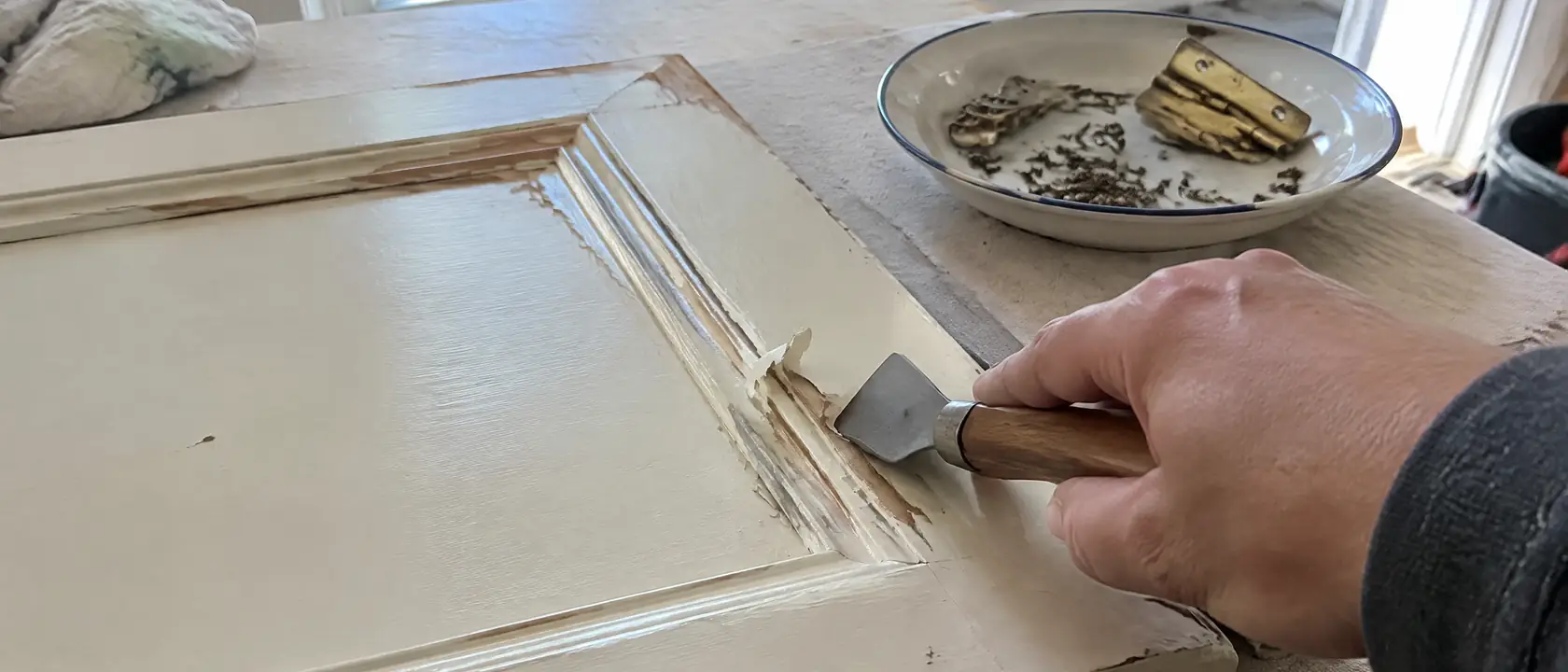 Hand scraping old paint off the molded edge of a period cabinet door, its original latches and hinges set aside in a dish