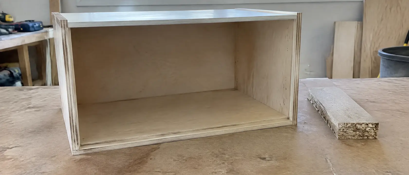 Cabinet box on its side showing the layered plywood edge and grooved bottom joint beside a crumbly particleboard offcut