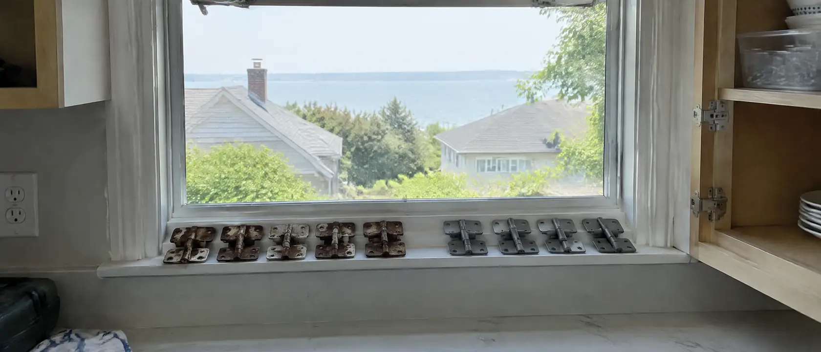 Rusted cabinet hinges on an open kitchen window sill beside new corrosion-resistant hinges, gray water past the rooftops
