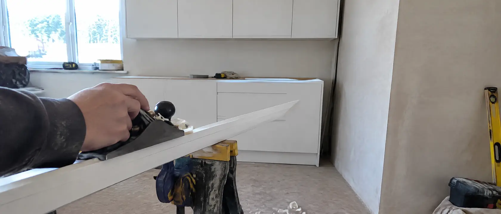 Hand planing a tapered filler strip to a pencil line, catalog wall cabinets behind stopping short of a leaning plaster wall