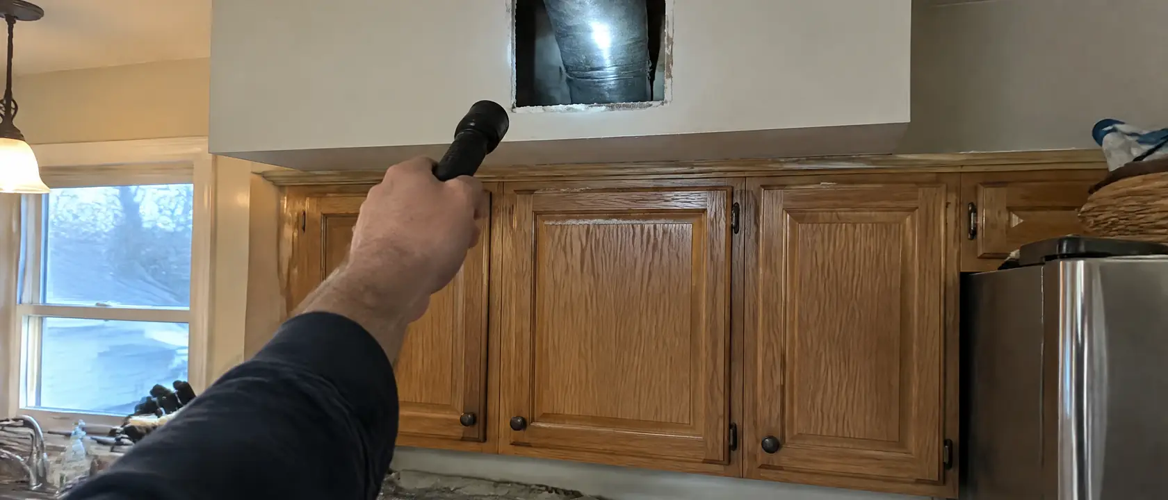 Inspection hole cut into a kitchen soffit above wall cabinets, a flashlight showing a metal duct inside the cavity