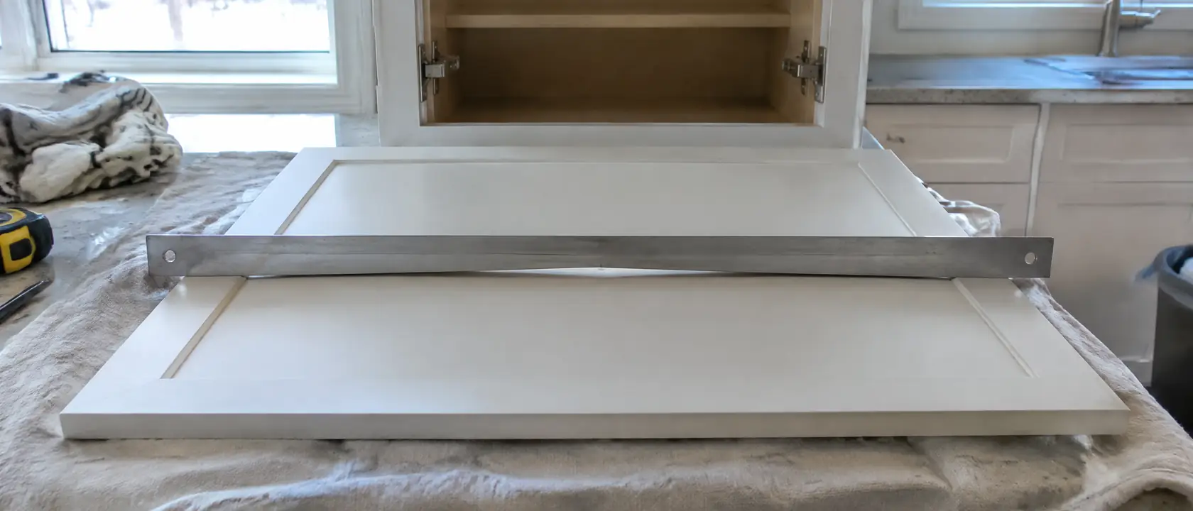 Straightedge laid across a cupped cabinet door on a drop cloth, a sliver of light showing under the middle