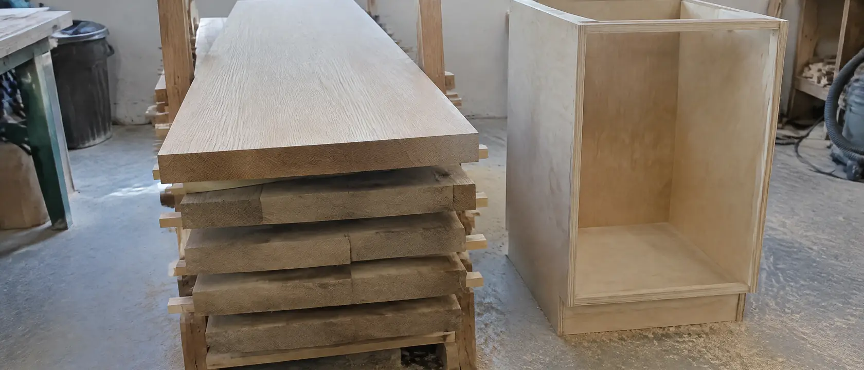 Rough quarter-sawn white oak boards on a shop rack, the top one planed clean, beside a plywood box awaiting its oak door