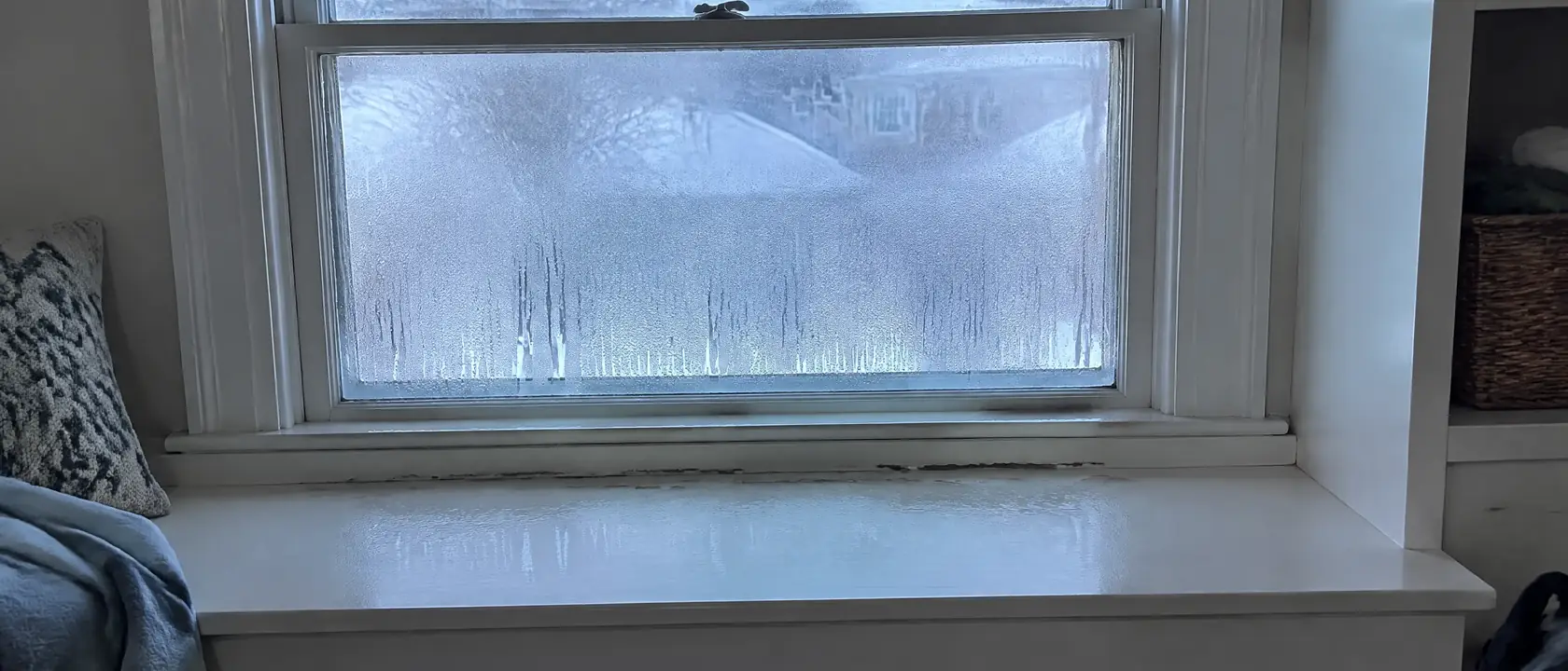 Condensation running down an old window onto the sill and pooling along the back edge of a painted window seat